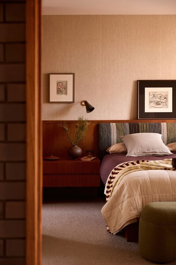 A warmly lit bedroom viewed through a timber-framed opening, featuring a richly stained wood bedside cabinet and wall wainscoting, textured beige wallcovering, framed artwork, a striped upholstered headboard, layered neutral bedding, and a rounded upholstered ottoman. The room has a cosy, refined mid-century-inspired feel with deep timber and muted earthy tones.