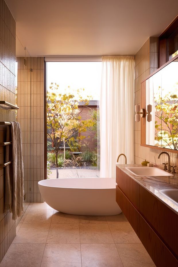 Warm contemporary bathroom with a freestanding white bathtub positioned beside full-height glazing and sheer curtains. The room features vertically gridded tan wall tiles, large beige stone-look floor tiles, a long timber vanity with integrated basins and dark metal taps, open shelving, and a large framed mirror. A landscaped courtyard is visible through the window and reflected in the mirror.