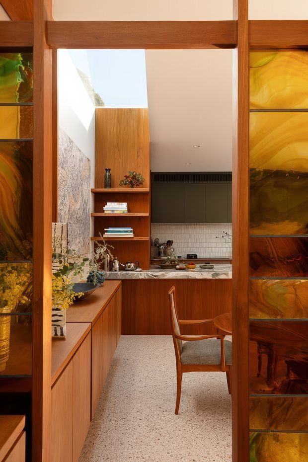A warm, compact mid-century-inspired kitchen viewed through a timber-framed opening with amber decorative glass panels. The space features extensive built-in timber cabinetry and shelving, a terrazzo-like light speckled floor, a white tiled splashback, dark upper cabinetry, stone benchtops, and a skylit or clerestory-lit area. A wooden dining chair and styled shelving add residential character.