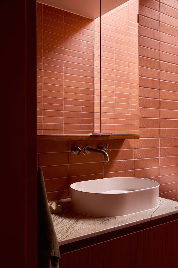 A compact bathroom vanity vignette with a round white vessel basin on a pale stone benchtop, wall-mounted brushed metal tapware, and a mirrored cabinet. The walls are clad in glossy terracotta-red rectangular tiles with matching grout, creating a warm, dramatic and richly monochromatic interior.