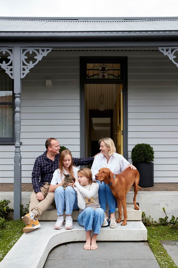 A family and dog posed on the front steps of a heritage-style house. The facade features pale horizontal weatherboard cladding, decorative timber fretwork and posts, a corrugated metal roof, dark-framed windows and entry door, and a small stained-glass panel above the doorway. A paved front path, raised garden beds and a potted topiary frame the entry.