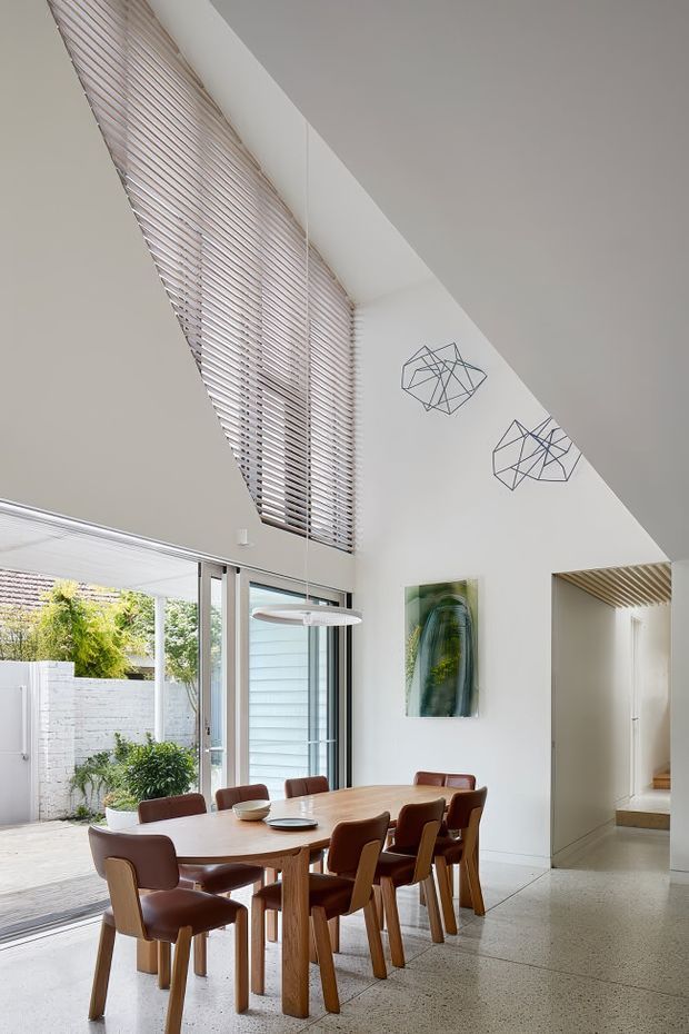 Bright contemporary dining area with a large timber dining table and upholstered brown dining chairs, positioned beside expansive glazed doors opening to a landscaped courtyard. The room has a high sloping ceiling, extensive horizontal blinds, polished light terrazzo or concrete flooring, white walls, a linear pendant light, abstract wire wall sculptures, and a stair or hallway opening with timber slatted ceiling detail.
