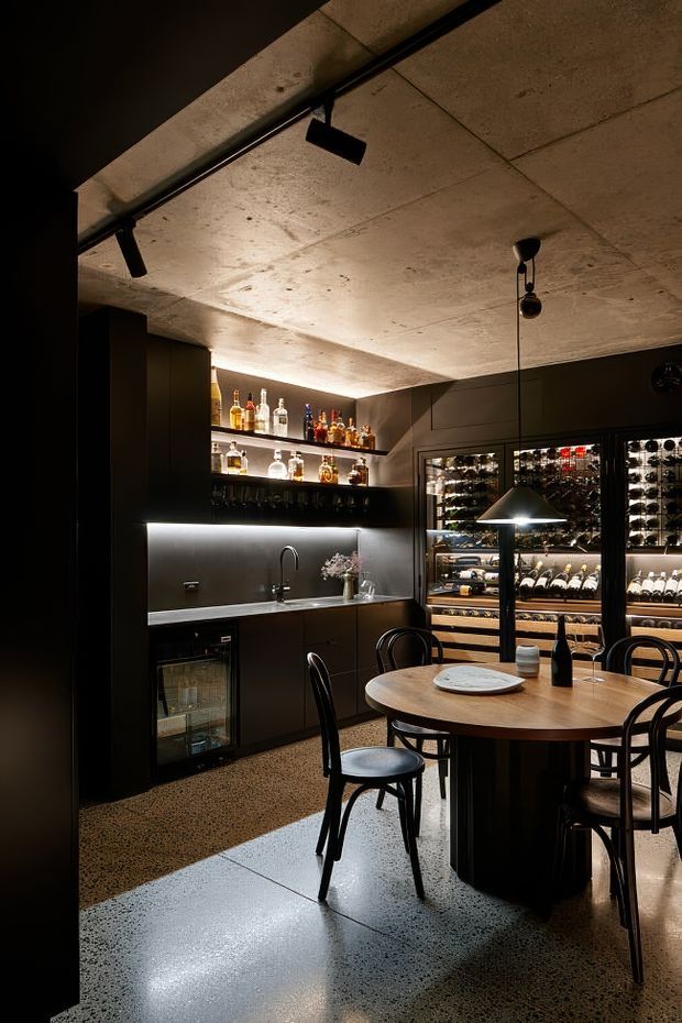 A compact, moody home bar and wine-storage area with dark built-in cabinetry, illuminated bottle shelving, a bar sink and under-counter beverage refrigerator. Glass-front wine fridges line the rear wall, while a round timber dining table with black bentwood chairs sits beneath a pendant light. The space features a raw concrete ceiling, polished speckled concrete floor and low, warm accent lighting.