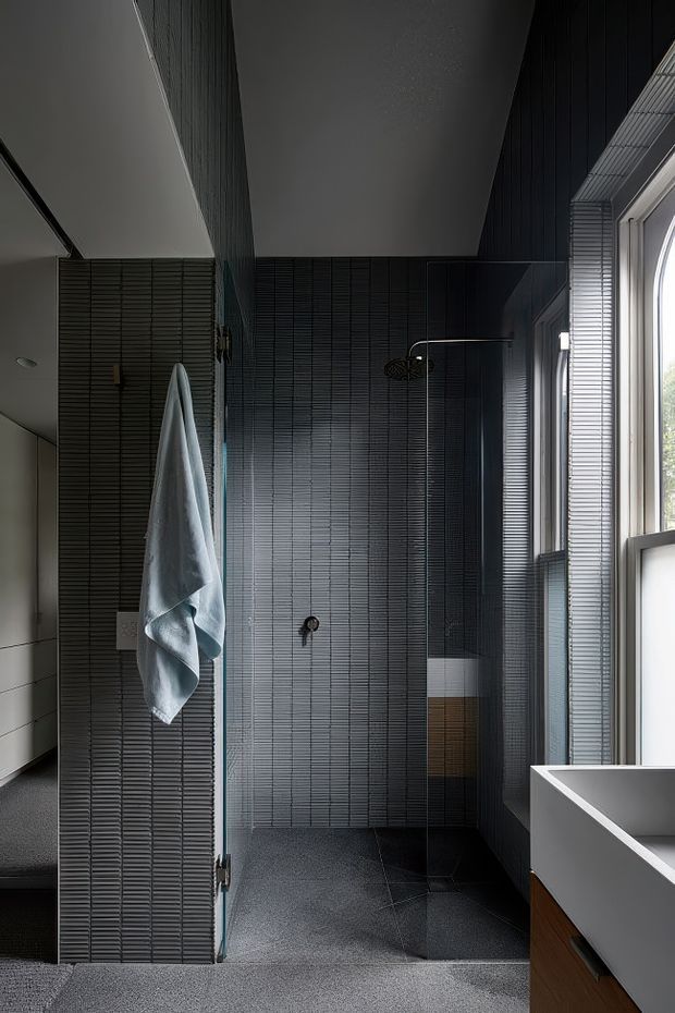 A dark, contemporary bathroom with full-height charcoal linear tile walls, matching dark floor tiles, a frameless glass shower enclosure, wall-mounted black shower fittings, and a freestanding or built-in white bathtub with timber-toned vanity cabinetry in the foreground. Tall windows bring in natural light, while a pale towel hangs beside the shower.