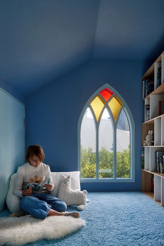 A cozy blue children's reading room with blue walls, ceiling, and carpet. A child sits reading on a low upholstered chair beside a small plush toy, with a built-in timber and white open bookshelf along the right wall. The room features a tall pointed-arch window with decorative stained-glass panels in red, yellow, and amber, bringing in natural light. The space has a calm, whimsical, storybook atmosphere.