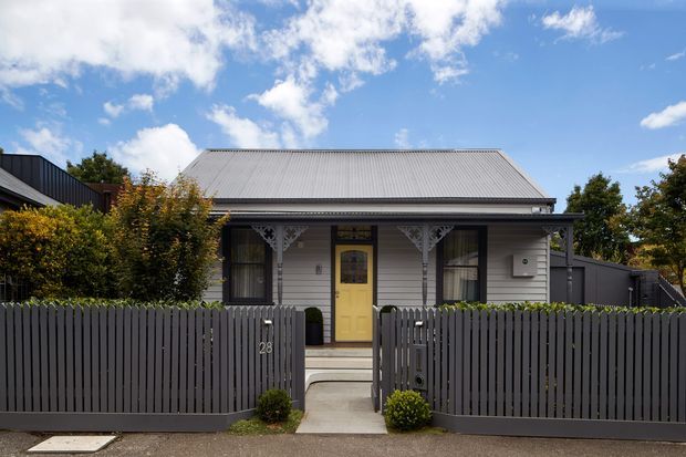 Front exterior view of a compact single-storey cottage-style house. The facade has pale horizontal weatherboard cladding, a low-pitched corrugated metal gable roof, dark-framed windows, decorative white fretwork brackets, and a yellow front door. A dark vertical-slat fence and landscaped shrubs enclose the narrow entry path.