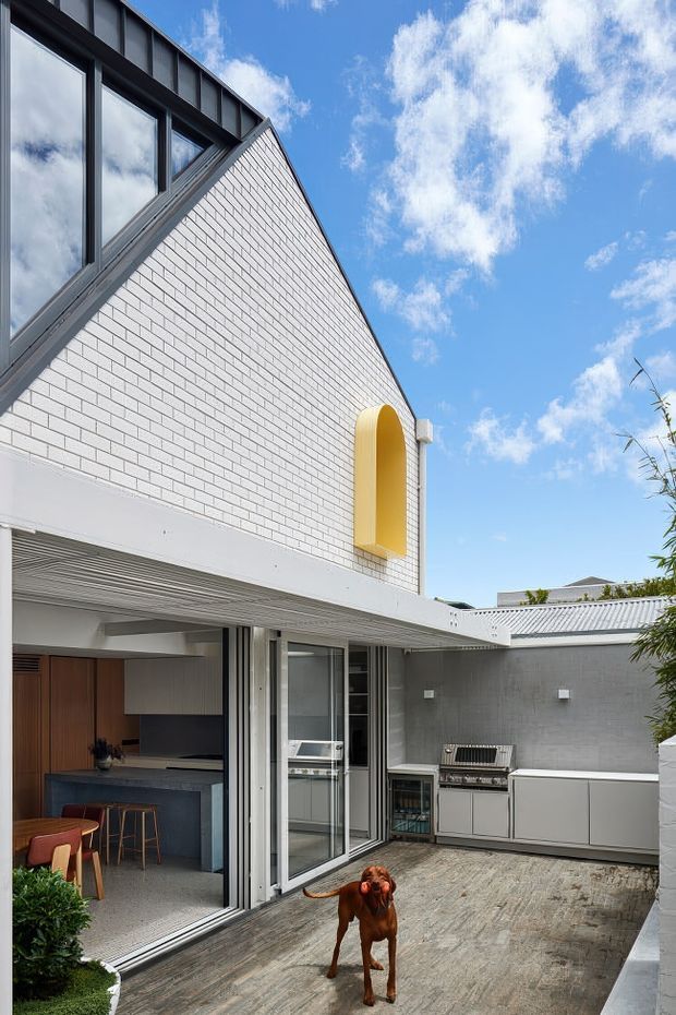 Exterior view of a contemporary courtyard house under a bright blue sky. The building combines white-painted brick walls, dark metal-framed glazing, a steep gabled upper volume, and a yellow arched wall recess. Large sliding glass doors open to an outdoor entertaining area with a built-in kitchen, barbecue, pale cabinetry, and textured paving. A dog is present in the courtyard.