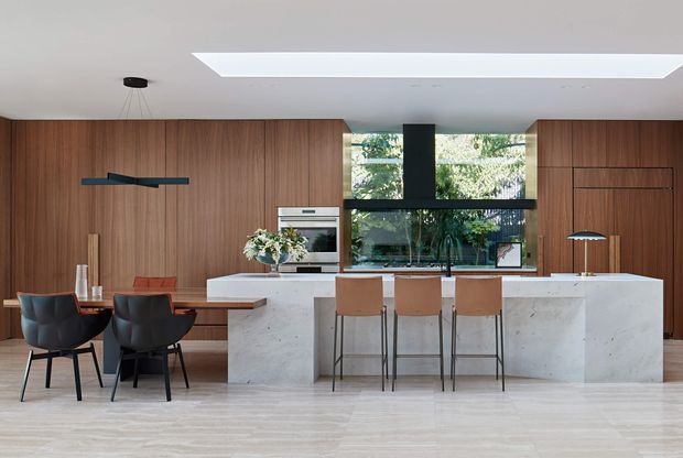 Contemporary open-plan kitchen and dining area with extensive warm timber wall panelling, a large pale stone island with integrated seating, built-in appliances, and a dark-framed glazed window overlooking greenery. A large linear skylight brightens the room, while a compact dining table and sculptural pendant add definition to the space.