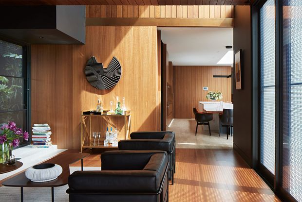 A warm, wood-lined contemporary living room with black leather lounge chairs, a low coffee table, drinks console, and sculptural wall art. Full-height glazing with blinds brings in strong sunlight and views of greenery. A dining area with a table, pendant light, and cabinetry is visible beyond the open passage, while exposed timber ceiling beams add architectural character.