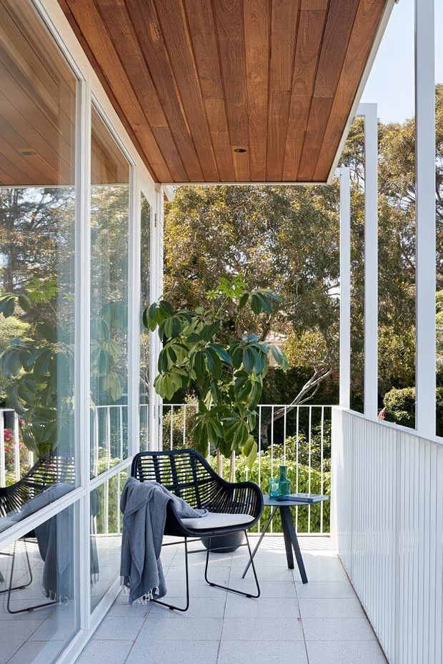 A bright, narrow enclosed balcony or terrace beside a glazed building facade. The space has white-framed floor-to-ceiling glazing, white vertical wall cladding, a white balustrade, pale tiled flooring, and a warm timber-lined soffit with a recessed light. A black woven lounge chair with a draped throw sits beside a small side table, overlooking dense leafy vegetation.