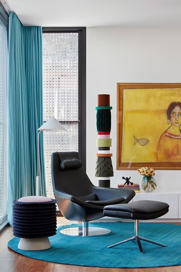 A bright, carefully styled living room corner featuring a black leather lounge chair with matching ottoman, a sculptural floor lamp, a round black side stool, vivid turquoise curtains and a large turquoise rug. A tall patterned window provides filtered light, while a colorful framed artwork and decorative stacked sculpture add visual interest against the white wall. Timber flooring is visible around the rug.