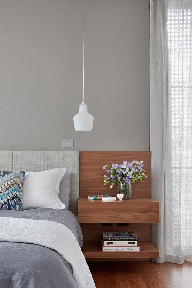 A calm, refined bedroom vignette featuring a grey upholstered bed with layered white and grey bedding, a warm timber floating bedside cabinet and open shelf, a white pendant light, and sheer floor-to-ceiling curtains beside the window. A vase of flowers and books add small decorative accents.