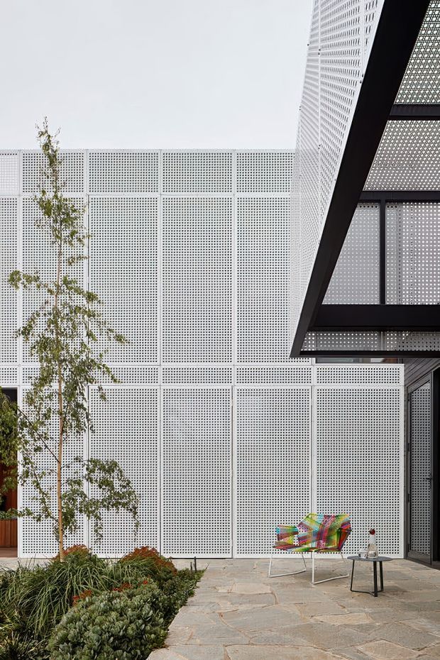 A contemporary outdoor courtyard or terrace with pale stone paving, layered low planting, and a sculptural multicoloured lounge chair. The space is enclosed by large white perforated screening panels, with a dramatic dark steel-framed canopy or overhang at the right. The image emphasizes geometric facade textures, filtered light, and the contrast between crisp architecture and soft landscaping.