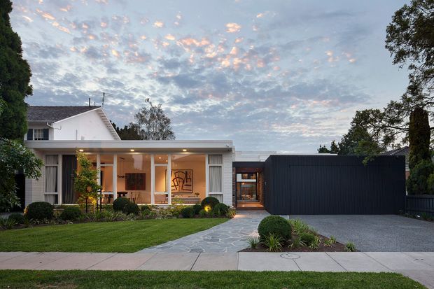 A low, contemporary single-storey home photographed from the street at dusk. The front facade combines a light rendered and glazed living wing with a dark vertically ribbed garage or secondary volume. A broad paved driveway, landscaped front garden, stone pedestrian path, mature trees and a covered entry are visible. A taller gabled residential form is set behind the modern flat-roofed addition.