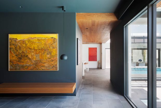 A refined contemporary interior hallway or entry space with a dark blue-grey feature wall, built-in timber bench, abstract artwork, and a black spherical pendant light. Large black-framed glazing on the right overlooks a bright outdoor pool area. The corridor continues toward a light-filled room with a timber-lined ceiling, pale tiled flooring, and a red artwork accent.