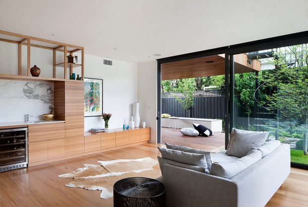 Bright contemporary living room with warm timber flooring and built-in timber cabinetry, an open shelving and media unit, a pale stone feature backsplash, and a large light-grey sectional sofa. Full-height black-framed glazing opens onto a timber deck and leafy outdoor area, creating a strong indoor-outdoor connection. A cowhide rug and restrained furnishings add texture while keeping the room airy and refined.