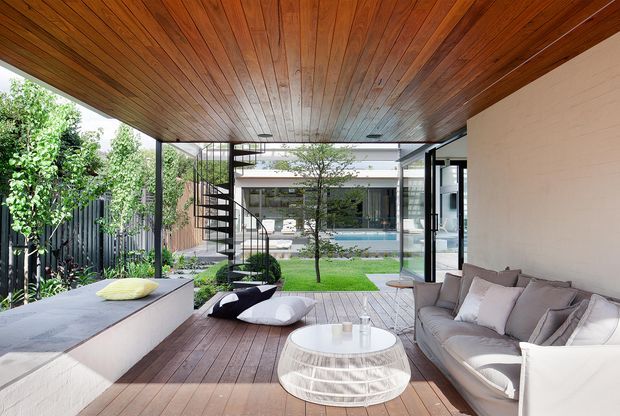 A refined covered outdoor living area with a timber-lined soffit and timber decking, furnished with a large upholstered sofa, a round white coffee table and floor cushions. The space opens onto a landscaped courtyard with a swimming pool and a modern residence beyond. A slim black spiral staircase and extensive glazing connect the outdoor area to the house.