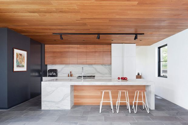 Contemporary kitchen with a large marble-look island and waterfall end, warm timber cabinetry and ceiling, dark charcoal feature wall, white integrated cabinetry, track lighting, and grey stone-look flooring. Three slim white bar stools sit at the island, with a large window providing natural light.