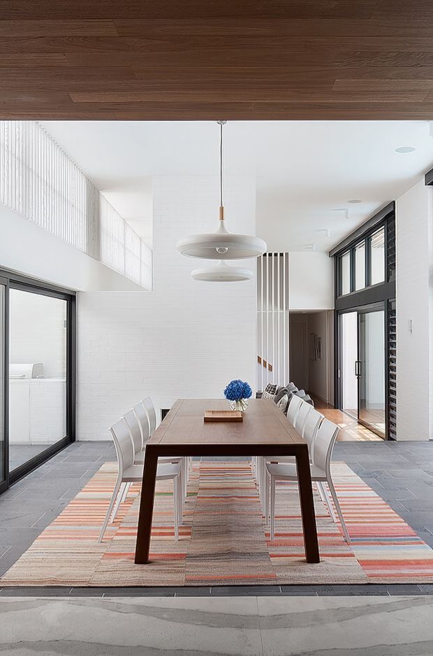 A bright, spacious dining area with a long dark timber dining table surrounded by white chairs. The room features white painted masonry walls, a high white ceiling with suspended pendant lights, expansive black-framed glazing and doors, grey tiled flooring, and a large striped area rug. A dark timber ceiling panel adds warmth and contrast, while a glimpse of a stairway and adjacent circulation space is visible in the background.
