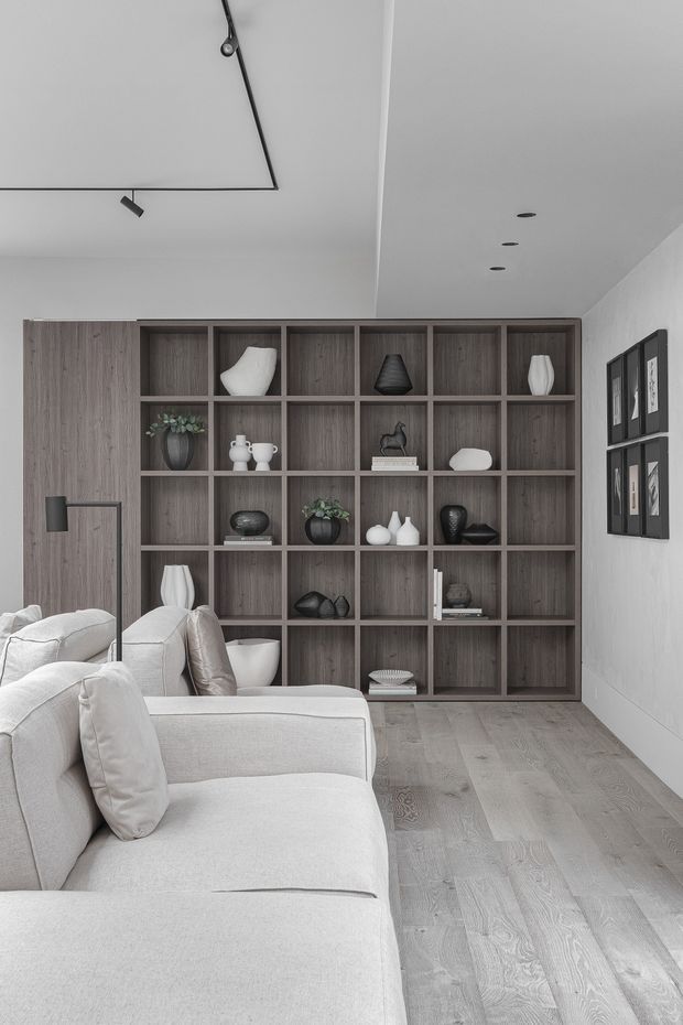 A refined contemporary living room with a large built-in dark timber display shelving wall, pale upholstered sectional seating, light timber flooring, framed artwork, and minimalist black track and recessed lighting. The restrained grey, white, and wood palette creates a calm, understated atmosphere.