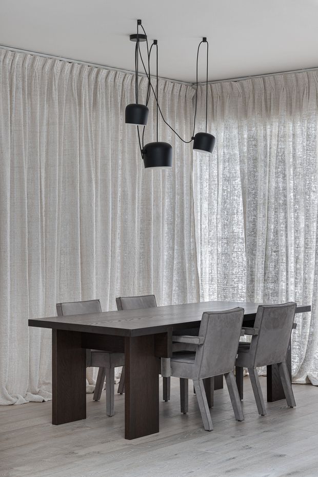 A refined dining room with a dark rectangular timber dining table surrounded by upholstered grey dining chairs. Floor-to-ceiling sheer textured curtains wrap around the corner windows, creating a soft, light-filled backdrop. Three black sculptural pendant lights hang above the table, and the pale timber flooring and neutral palette give the room a calm, contemporary atmosphere.
