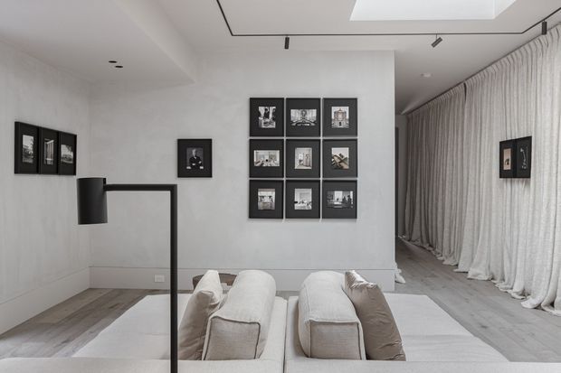A refined monochromatic living room with pale textured walls, light timber flooring, sculptural cream sofas, and a black minimalist floor lamp. Black-framed photographic artwork forms gallery displays on the walls, while full-height sheer curtains line the right side. A recessed skylight and slim black track lighting add architectural interest.