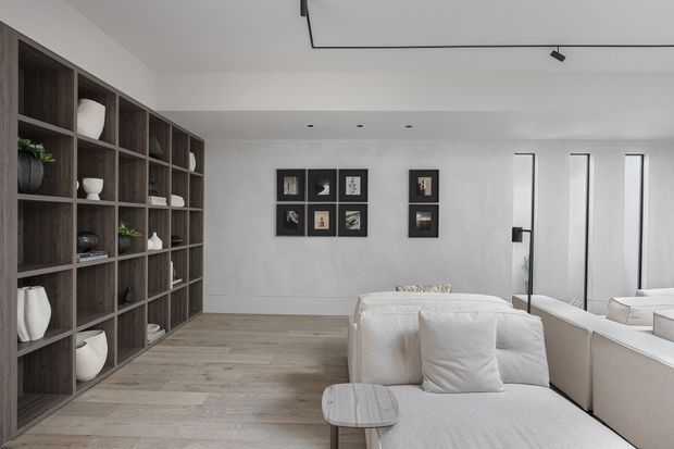 A bright, refined contemporary living room with a large built-in dark timber display shelving unit along the left wall, pale timber flooring, white textured walls, framed artwork, and a light upholstered sectional sofa. Full-height mirrored panels add depth on the right, while slim black track lighting provides a restrained modern detail.