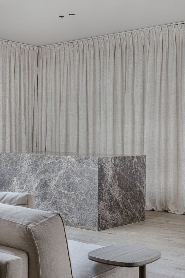 A refined contemporary living room featuring full-height textured cream curtains, pale timber flooring, a large veined grey marble-clad fireplace or feature block, and a neutral upholstered sofa with a rounded stone or timber coffee table. The restrained palette and soft textures create a calm, luxurious atmosphere.