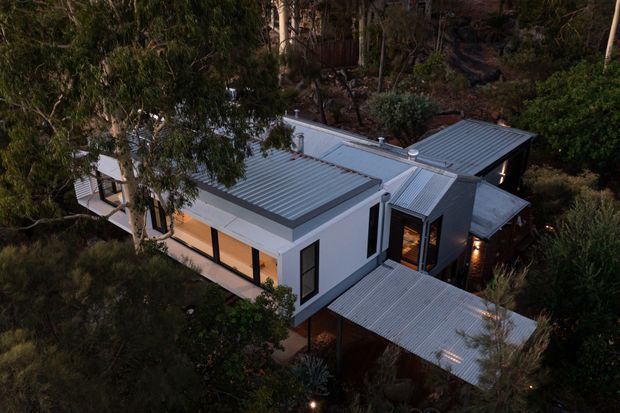 Aerial dusk view of a modern single-storey home set within dense native vegetation. The building has a fragmented low-profile form with multiple corrugated metal roof planes, pale rendered or panelled walls, dark-framed glazing, and a covered outdoor terrace or entry canopy. Warm interior lighting contrasts with the cool grey roof and surrounding bushland.