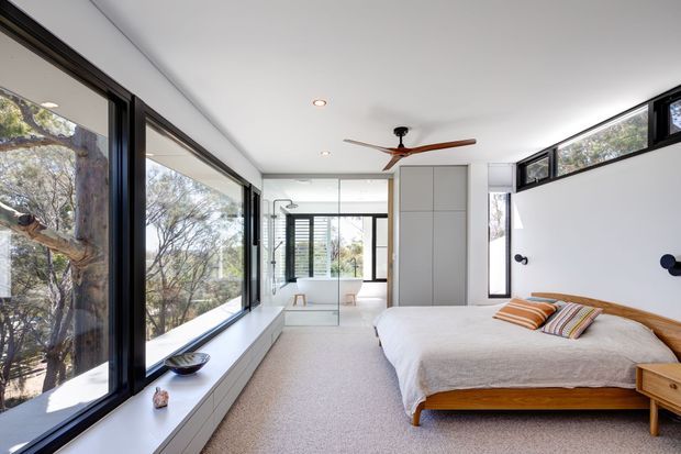 Bright contemporary bedroom with expansive black-framed glazing overlooking trees, a low built-in window seat and storage joinery, white walls, and a timber platform bed. A glass-enclosed bathroom is visible beyond the bedroom, with a ceiling fan and restrained striped and rust-toned bedding adding warmth.