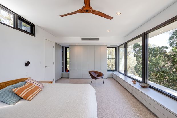 A bright, contemporary bedroom with a low-profile bed, white bedding, and a sculptural leather lounge chair. The room features extensive floor-to-ceiling corner glazing with views of trees, integrated white cabinetry beneath the windows, a built-in wardrobe or storage wall, recessed ceiling lights, and a timber ceiling fan. The palette is predominantly white and light neutral with warm timber and tan accents.
