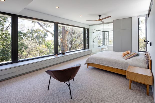 Bright contemporary bedroom with expansive black-framed corner windows overlooking trees, a low timber bed, grey bedding, integrated light-grey wall cabinetry, carpeted floor, and a sculptural leather lounge chair. A mirrored wall reflects the glazing and room, while recessed ceiling lights and a ceiling fan provide understated illumination and ventilation.
