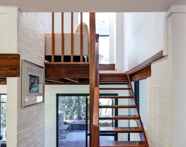 Interior view of a compact loft-like stairway and landing with open-riser timber stairs, dark timber handrails and balustrades, white-painted brick walls, and a partially visible upper-level bedroom or sleeping platform. Glazing and a lower doorway provide views to an outdoor area, while framed artwork and exposed timber structural elements add warmth. The composition emphasizes vertical circulation and the mezzanine connection.