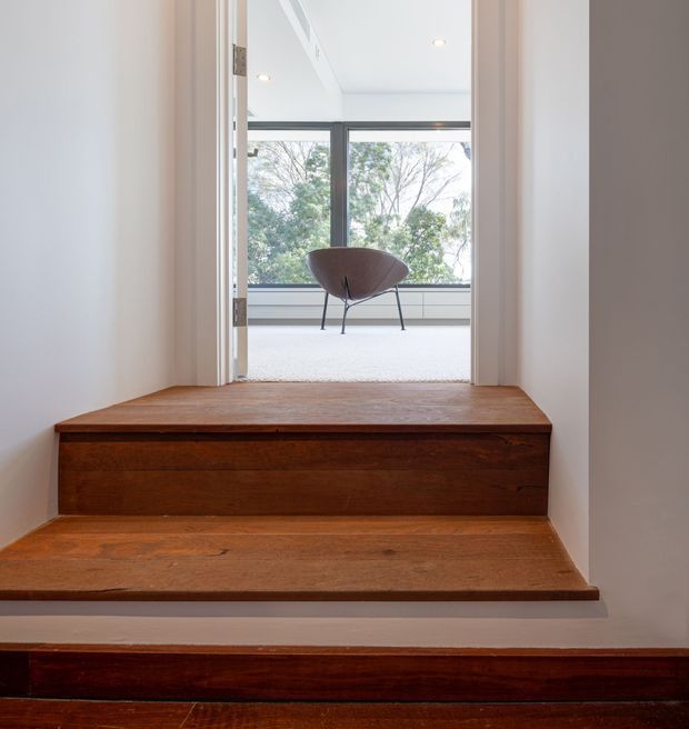 Interior view looking up a short set of broad timber steps toward a bright room. The stair treads, riser, and lower skirting are dark warm wood, contrasted with smooth white walls and a white ceiling. A partially open white door frames the room beyond, which has large dark-framed windows overlooking leafy trees and a sculptural mid-century-style lounge chair. The space is simple, light-filled, and restrained.
