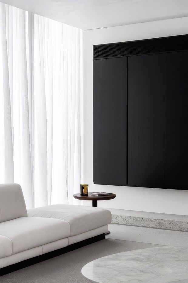 A minimalist living room with a large white upholstered chaise sofa, sheer floor-to-ceiling curtains, a black wall-mounted entertainment or feature panel, and a small round side table. The room uses a restrained monochromatic palette with pale flooring and a sculptural light stone or terrazzo hearth-like platform.