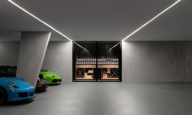 A sleek, dark-toned luxury garage with polished grey concrete surfaces, concealed linear LED lighting, angled structural wall elements, and a glass-fronted wine storage display at the rear. Several brightly coloured sports cars are parked along the left side, creating a high-end automotive showroom atmosphere.