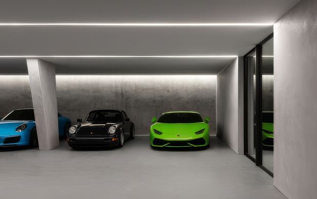 A sleek, minimalist private car garage with three sports cars displayed against a dark textured concrete wall. The space features a smooth grey floor, white structural columns, full-height dark-framed glazing or doors on the right, and continuous recessed linear ceiling lighting.