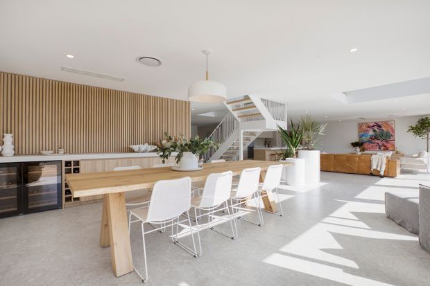 Bright, open-plan contemporary dining and living area with a long solid timber dining table and white wire-frame chairs in the foreground. A vertical timber slat feature wall runs behind a built-in bar and wine fridge, while a white open staircase with timber treads leads upward. The space has a pale polished concrete floor, white ceiling with recessed lighting and a linear air vent, large skylight-like openings, indoor plants, and warm timber cabinetry. A living area with a grey sofa and colourful artwork is visible to the right.