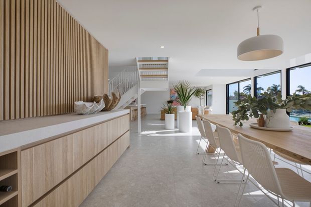 Bright open-plan contemporary interior combining a dining area and kitchen joinery. A long light timber dining table with white chairs sits beside expansive glazing, while a pale timber built-in sideboard and vertical timber batten feature wall define the left side. The space has a white ceiling, polished light grey floor, sculptural pendant light, indoor plants, and a staircase leading to an upper level.