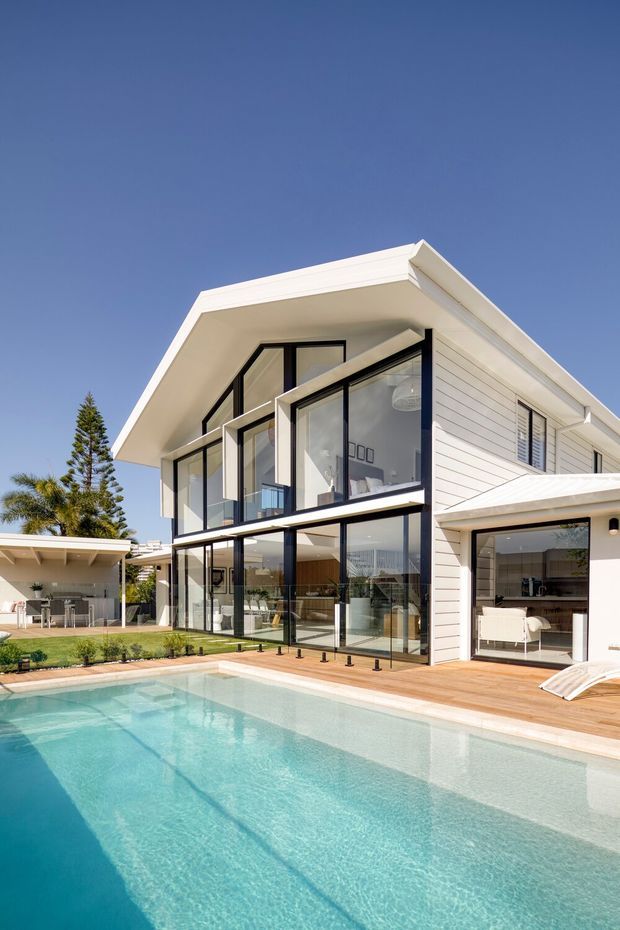 Bright exterior view of a large contemporary two-storey house beside a swimming pool. The home features white horizontal weatherboard cladding, a pitched gable roof with broad eaves, extensive black-framed floor-to-ceiling glazing, and a timber pool deck. A covered outdoor entertaining area and an open-plan interior are visible through the glazing, with landscaped planting surrounding the pool.