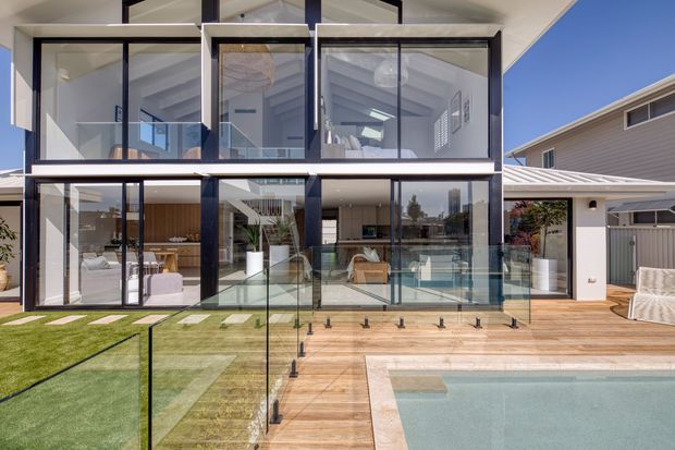 Contemporary multi-storey residence viewed from the backyard, featuring extensive floor-to-ceiling glazing, dark framed windows, a timber-clad terrace and pool deck, frameless glass pool fencing, and a swimming pool. Bright interiors and open-plan living spaces are visible through the glazing, with a neighbouring suburban house at right.