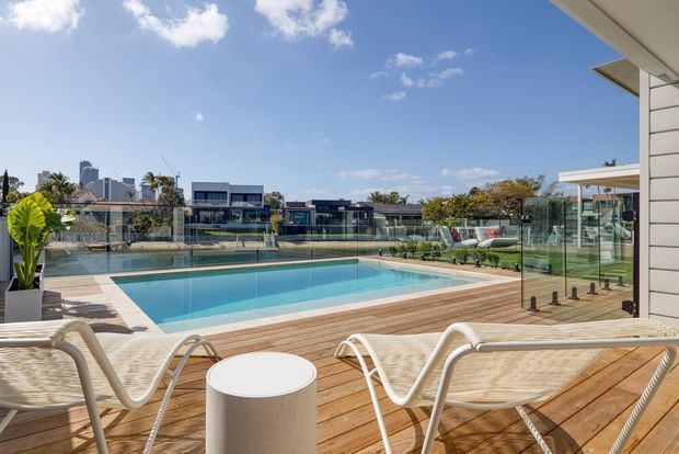 A bright contemporary outdoor pool area viewed from a timber deck, with a rectangular swimming pool enclosed by clear glass balustrades. White lounge chairs and a small round side table occupy the foreground, while a modern low-rise residential neighbourhood and distant city skyline are visible beyond. The scene has a clean, refined resort-like atmosphere with strong indoor-outdoor connection.