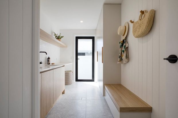 Bright, narrow contemporary entry and utility corridor with pale timber built-in cabinetry, a long bench seat, white vertical wall panelling, light tiled flooring, and a compact kitchenette or laundry zone along the left wall. A black-framed glazed door provides light at the far end, while woven baskets, hats, and hanging textiles add relaxed coastal character.