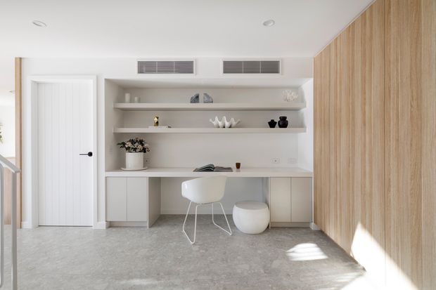 Bright minimalist home office or study nook with a built-in wall desk, open display shelving, white furniture, pale timber wall panelling, and a polished grey concrete-like floor. A white internal door and concealed air-conditioning vents are visible, with strong natural light entering from the right.