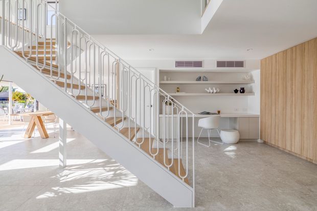 Bright contemporary open-plan interior featuring a sculptural white staircase with slender looping metal balustrades, pale stone or terrazzo flooring, a built-in study nook with open shelving and desk, and warm vertical timber wall panelling. Sunlight enters from the left, creating an airy, refined atmosphere.