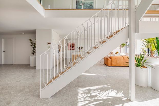 Bright, open-plan interior dominated by a sculptural white staircase with decorative looped metal balustrades. The space has pale stone or concrete-look flooring, white walls and structural columns, an upper-level landing, indoor plants, and a glimpse of a tan sofa in the living area beyond. Strong daylight creates an airy, refined atmosphere.
