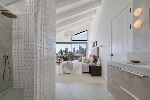 A bright contemporary ensuite bathroom opening directly into a bedroom. The bathroom features a walk-in shower with small white rectangular wall tiles, a frameless glass partition, a pale stone-look floor, floating light-wood vanity cabinetry, and a white basin. The adjoining bedroom has a pitched white ceiling with exposed beams, soft neutral bedding, and large windows overlooking a dense city skyline. Rounded wall sconces and restrained neutral furnishings create a calm, refined atmosphere.