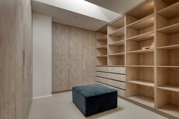 A custom walk-in wardrobe with full-height light timber cabinetry, integrated hanging storage, open shelving, and a central bank of drawers. A dark upholstered ottoman sits on a pale carpeted floor. The room is softly lit by a large ceiling skylight or rooflight, creating a clean, warm, minimalist storage space.