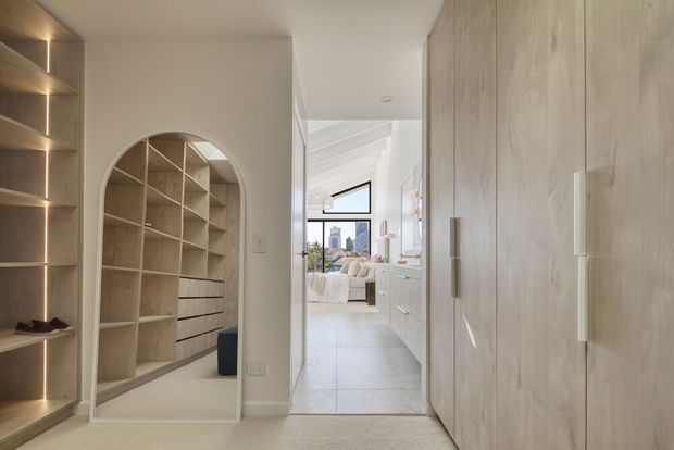 A bright, refined interior hallway or dressing area with extensive pale timber built-in shelving and cabinetry. The foreground features tall closed wardrobe doors on the right and illuminated open shelving on the left. An arched opening reveals additional timber shelving and drawers, while a bedroom with white bedding and large windows is visible beyond. The space has light neutral walls, pale tiled flooring in the adjoining room, integrated lighting, and a clean contemporary fit-out.