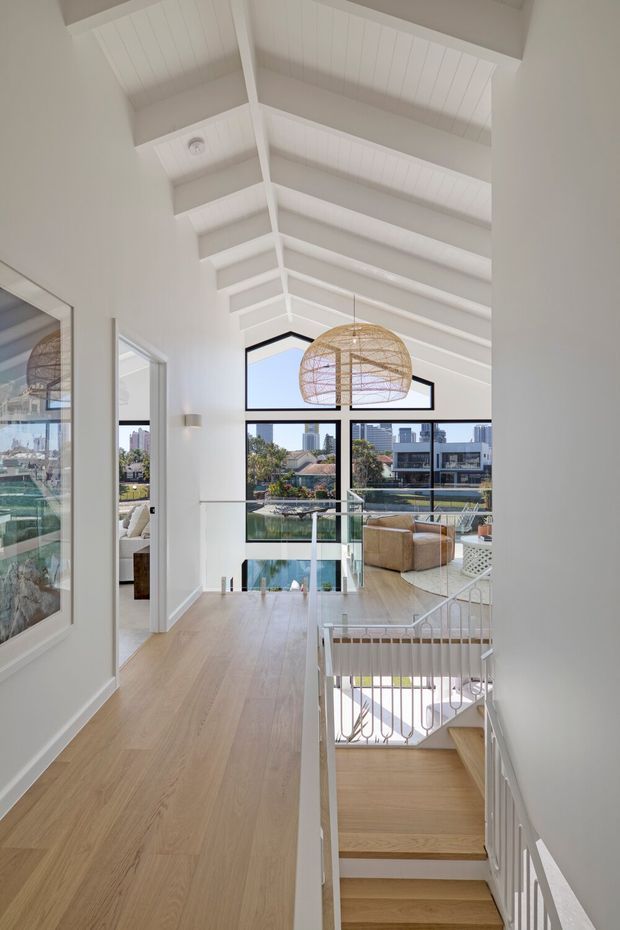 A bright, refined residential hallway and stair landing overlooking an open-plan living area. The space features a high white vaulted ceiling with exposed painted beams and panelled lining, expansive black-framed windows, pale timber flooring, glass balustrades, and a white metal stair railing. A woven pendant light hangs above the living room, with a cityscape visible beyond the glazing.